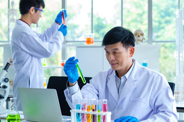 Portrait shot of Asian professional mature male scientist in white lab coat and rubber gloves sitting look at camera smiling holding blue liquid sample in test tube while collegaue working behind