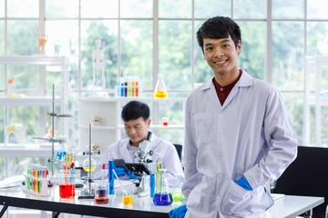 Portrait shot of Asian professional successful young handsome male scientist in white lab coat rubber gloves standing crossed arms smiling look at camera in laboratory while colleague working behind