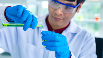 Closeup shot hands of Asian male scientist in white lab coat safety goggles and rubber gloves in blurred background careful holding pouring green sample from test tube to glass vial in laboratory