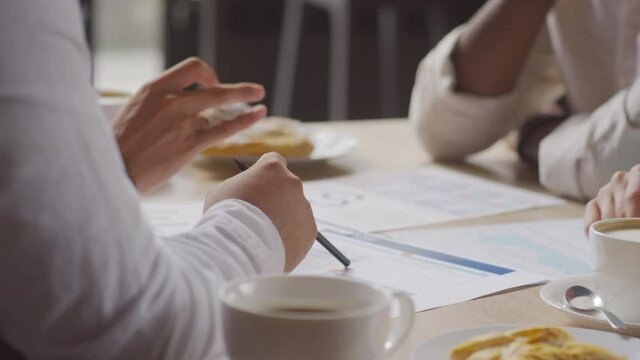 Low angle of hands of four unrecognizable colleagues sitting at table in cafe, having coffee, working with documents, having business meeting