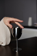 Shot of a female hand with a steel wine glass. The fingers are on the top of the metal glass. The goblet stands on a black table. There is a dark gray wall and different items at the background.
