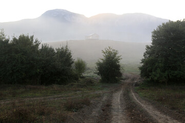 Light morning fog disappears with sunrise. Path in the field, trees and silhouette of mountains