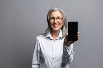 Promotion concept. Pleasant senior gray-haired business woman in glasses and formal wear holding and showing smartphone with empty screen standing on gray, mature lady advertising new app, sale deal - obrazy, fototapety, plakaty