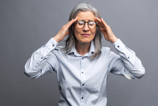 Tired senior mature gray-haired businesswoman holding forehead standing isolated on gray with eyes closed. Elder female entrepreneur ceo manager feels headache, suffers from migraine, burnout