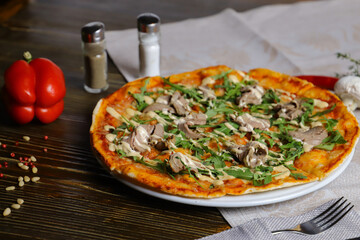 Meat mushroom pizza on a wooden table Nearby is a red bell pepper a salt shaker and a pepper shaker and cutlery.