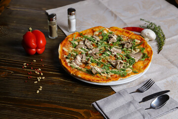Meat mushroom pizza on a wooden table Nearby is a red bell pepper a salt shaker and a pepper shaker and cutlery.