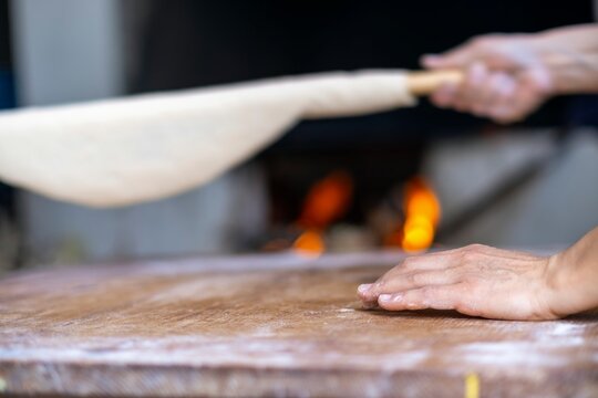 Female Hands Of Baker Rolls Dough In Flour On Wooden Board On Table With A Rolling Pin, In Bakery Shop, Woman Making Pizza Using Traditional Recipe At Home.
