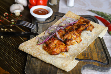 Close-up of a shish kebab on pita bread with rings of red onion on a cutting board on a linen napkin, in a ketchup gravy boat.