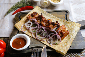 Close-up of a shish kebab on pita bread with rings of red onion on a cutting board on a linen napkin, in a ketchup gravy boat.