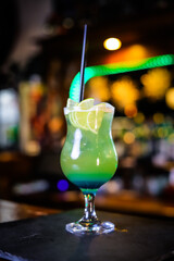 A cocktail in a tall green glass, lemon wedges and a black straw. Filming of the bar menu against the background of bright lights in bokeh.