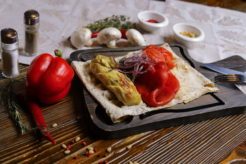 The pita bread is served with grilled vegetables zucchini, red pepper, a skinless tomato on a dark cutting board, a linen napkin on a dark table top. There is a couple of pepper and salt
