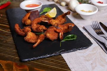 Shrimp is served with sauce. Tempura king prawns, sliced lemon, green basil leaves are served on a black stone stand. Cutlery, hot pepper pods, spices in bowls, white mushrooms.