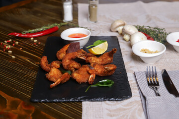 Shrimp is served with sauce. Tempura king prawns, sliced lemon, green basil leaves are served on a black stone stand. Cutlery, hot pepper pods, spices in bowls, white mushrooms.