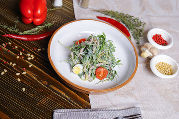 On a dark countertop, a light linen napkin, on a cutting board Served salad with rucola leaves, boiled tongue cut into long slices with sauce A couple of salt and pepper, cutlery, red peppers decorate