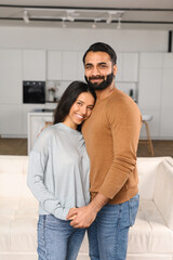 Smiling man hugging his charming woman. Two people standing and joyfully looking at the camera. Young Indian couple happily spending time in cozy modern kitchen at home