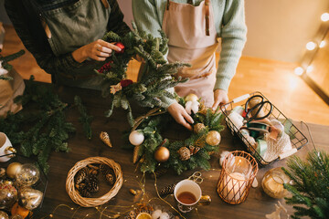 Two millennial women making Christmas wreath using pine branches and festive decorations. Small...
