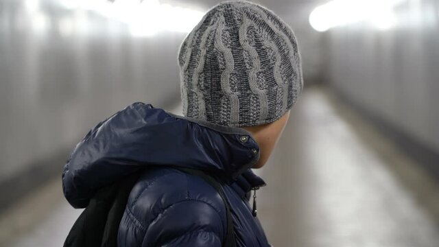 Portrait of a lonely sad boy in a hat and jacket with a backpack turned away from the camera in an underground covered passage in the city in winter