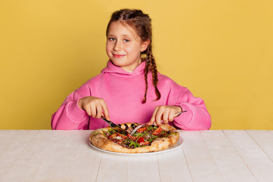 Studio shot of little cute girl sitting and tasting delicious Italian pizza isolated on yellow studio background.