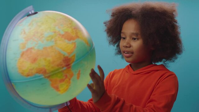 Curious African American school girl exploring the globe standing on blue background. Education process for kids.