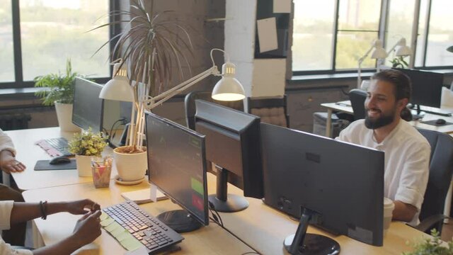 Arc of young Black and bearded Biracial man sitting at desks in front of each other, smiling and bumping fists over computers in open space office at daytime