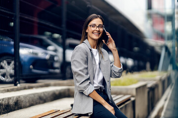 A happy young, elegant woman is sitting outdoors and having a phone call. A woman is sitting on a bench and having a phone call. A businesswoman talking on the phone outside