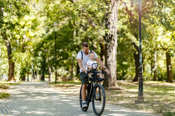 Obraz premium Active family vacation and match casual clothes. A male toddler and a father ride a bike around the park. Cycling holiday and active weekend. Toddler sits in the basket on the bike and laugh with dad