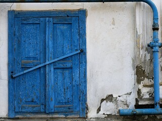 a fragment of the facade of an old destroyed building with white fallen off plaster and blue shutters of a closed window and a peeling blue gas pipe, a dilapidated abandoned building outside view