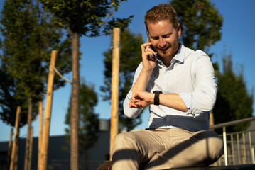 Young stylish businessman sitting on the beanch outdoors. Portrait of handsome man using the phone