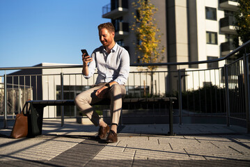 Young stylish businessman sitting on the beanch outdoors. Portrait of handsome man using the phone