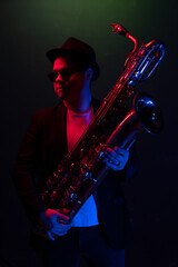 Young Saxophonist in a Black Jacket, Hat and Sunglasses Holds a Baritone Saxophone. Dark...