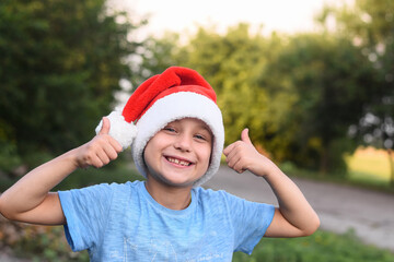 portrait of boy happy, smiling in red santa hat giving thumbs up sign with fingers. Positive emotion and symbols. outdoors against the background of green summer trees.