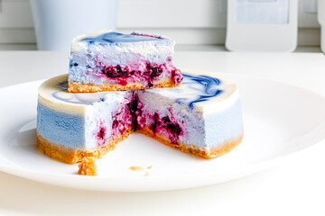 Piece of blue and white cheesecake with blueberry, cherry and black currant sauce inside on white plate on white table