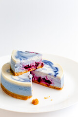 Piece of blue and white cheesecake with blueberry, cherry and black currant sauce inside on white plate on white table