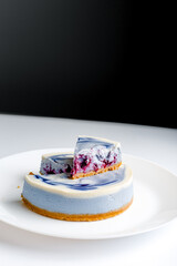 Piece of blue and white cheesecake with blueberry, cherry and black currant sauce inside on white plate on white table and dark black background
