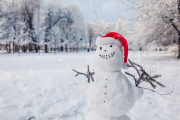 Happy snowman wearing Santa hat in snowy winter park at Christmas season outdoors. Winter activities