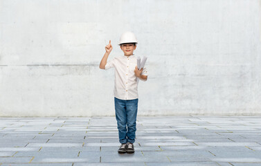 building, construction and profession concept - smiling little boy in helmet with blueprints pointing finger up over grey concrete wall background