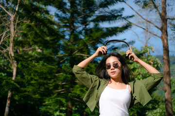 girl wearing headphone and sunglasses,enjoy listening music in the park,relax time,pine tree as background,nature outdoor park,green jacket and white shirt lady