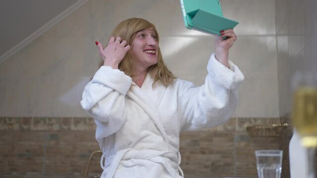 Smiling happy trans woman waving talking at tablet sitting in bathroom at home. Portrait of confident Caucasian LGBTQ person blogging indoors chatting online in social media using video chat
