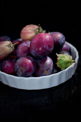 Seasonal Blue and purple Fruit and vegetable bowl with green leaves in a bowl on rustic dark background, top view. Plum and Organic Raw Baby Indian Eggplants