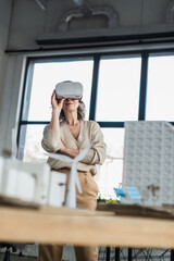 Mature businesswoman using vr headset near blurred models of buildings in office.