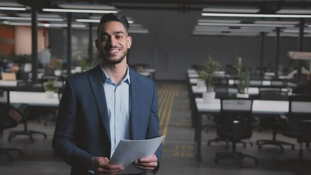 Young arab businessman in formal suit smiling to camera, posing at modern office interior, tracking shot, free space