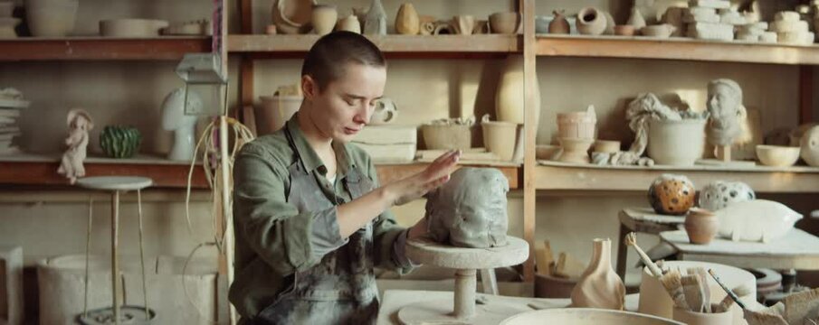 Zoom in shot of young female artist in apron sculpting clay head by hand in workshop