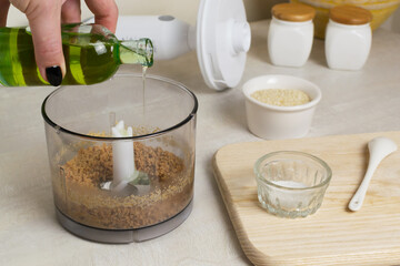 Woman pouring oil into sesame seeds in a blender bowl at home kitchen. The process of making a traditional tahini sauce. Levantine cuisine concept.