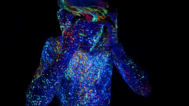 Man with headache from an overdose is standing in dark studio painted with fluorescent paints, and shows negative emotions, front view. Man painted body paints have badtrip