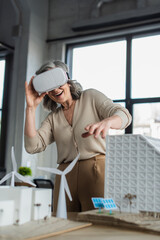 Happy businesswoman in virtual reality headset near models of buildings in office.