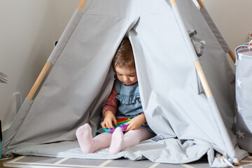 childhood, hygge and people concept - little baby girl playing with simple dimple toy in kid's tent or teepee at home - obrazy, fototapety, plakaty