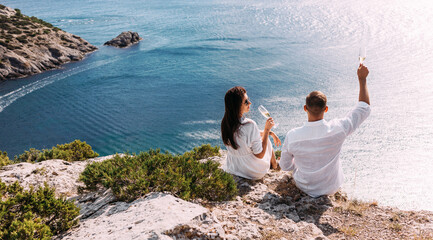 A man and a woman drink champagne on the seashore, rear view. Wedding trip to the sea, panorama. A couple in love in white clothes on the coast. Couple with champagne by the sea. Wedding travel