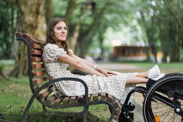 Pleasant young woman keeping her feet on wheelchair while sitting on wooden bench among green...