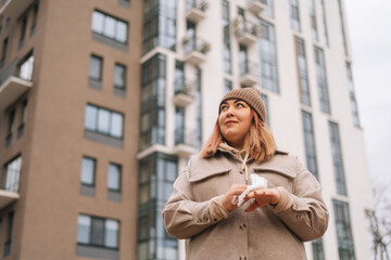 Fototapeta premium Shooting from below of overweight woman using antibacterial wet wipes to clean and disinfect hands outdoors, standing in urban street on blurred background on modern building in cloudy autumn day.