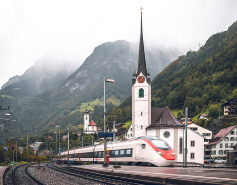 Scenic landscape of Swiss Alps in rainy weather: High-speed train of Swiss railways SBB arriving at Fl&uuml;elen station (Switzerland), passing church and mountains covered in fog.